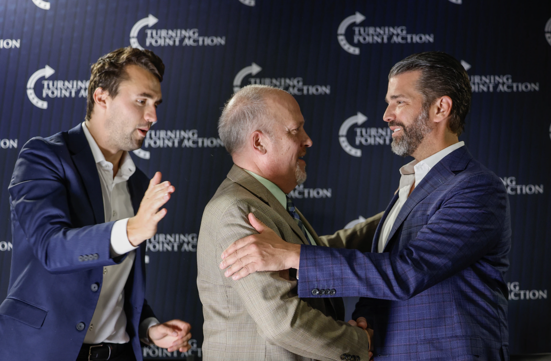 Schimel shaking hands with Donald Trump Jr. with Charlie Kirk about to tap him on the back.